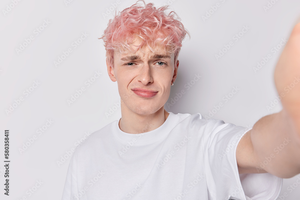 Displeased hipster guy with dyed pink hair has sulking gloomy expression dissatisfied with something dressed in casual t shirt keeps arm outstretched to make selfie isolated over white background Displeased hipster guy with dyed pink hair has sulking gloomy expression dissatisfied with something dressed in casual t shirt keeps arm outstretched to make selfie isolated over white background