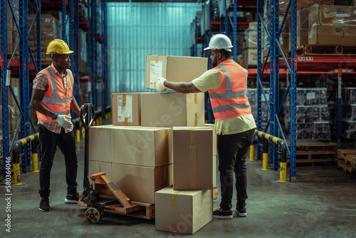 Two African American warehouse workers working together in warehouse moves cardboard boxes using manual pallet Truck.