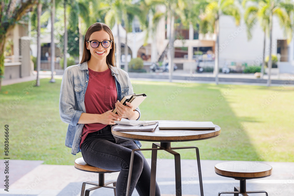 Young female student sitting on chair and desk and holding books and smiling in camera in college or university building. Young female student sitting on chair and desk and holding books and smiling in camera in college or university building.