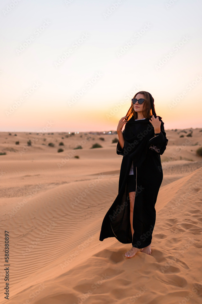 Beautiful mysterious woman in traditional arabic black long dress stands in the desert on sunset - obrazy, fototapety, plakaty 