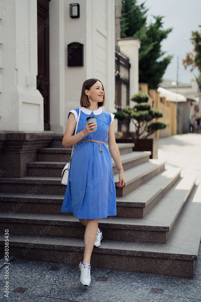 Fototapeta premium Beautiful young pregnant woman in a blue dress walking through the city streets having a take away drink.