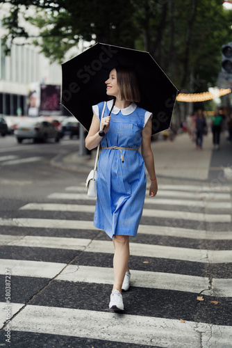 Pretty woman, in a blue dress, with a white purse and an umbrella passing the crosswalk