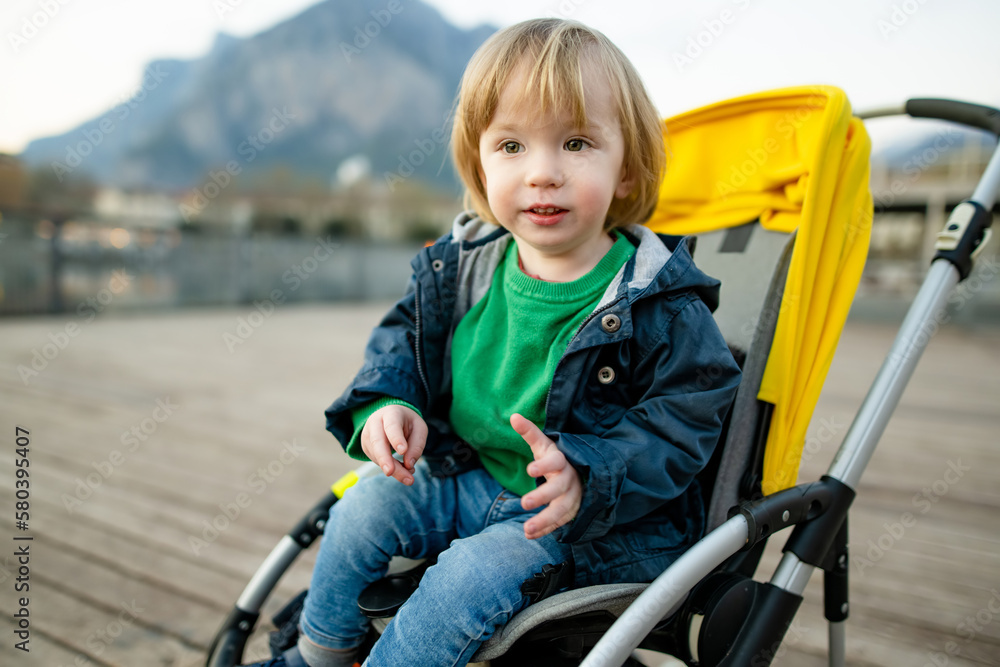 Sweet toddler boy sitting in a stroller in Lecco town. Little child in pram. City walks with kids. Sweet toddler boy sitting in a stroller in Lecco town. Little child in pram. City walks with kids.