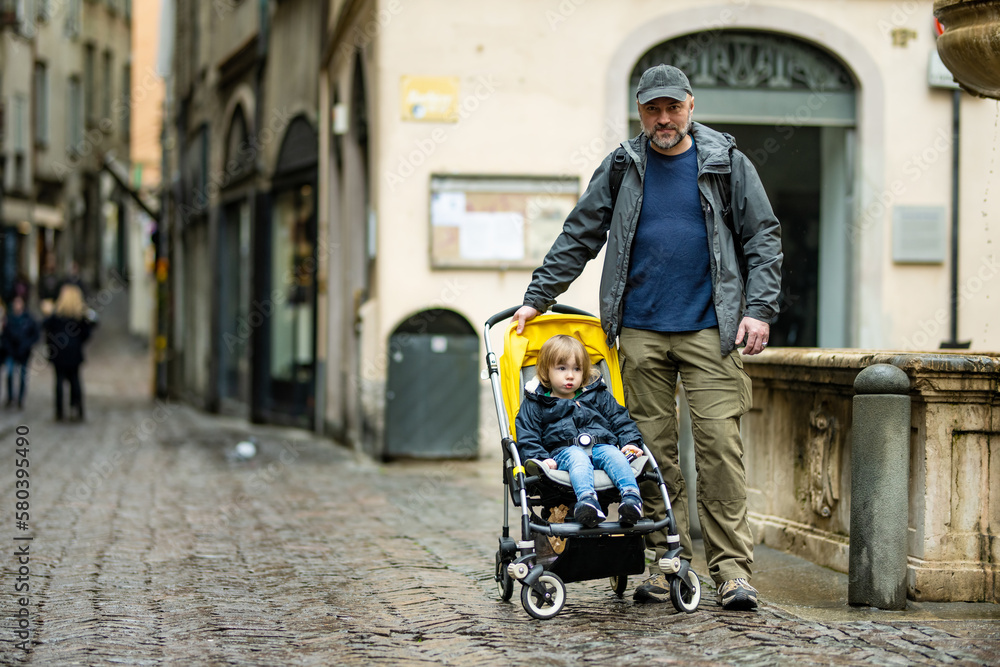 Obraz premium Young father and his toddler son in a stroller walking together down the medieval streets of Bergamo city northeast of Milan. Scenic views of Citta Alta, town's upper district.