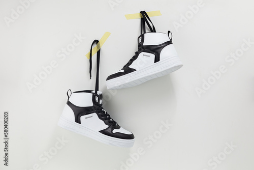 Pair of modern trendy sneakers hang on laces on color wall. White and black unisex sport footwear hanging on duct tape. Place for text. Creative minimal design. photo