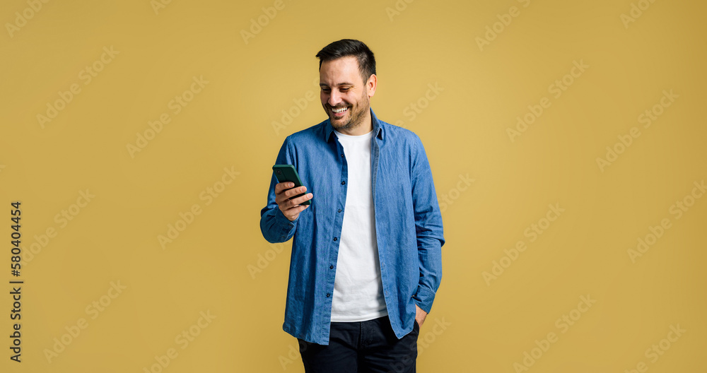 Smiling mid adult handsome man browsing social media on smart phone while posing with hand in pocket on isolated yellow background Smiling mid adult handsome man browsing social media on smart phone while posing with hand in pocket on isolated yellow background