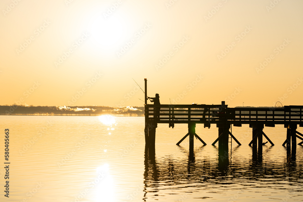 A silhouette of a person fishing off of a pier during sunrise.