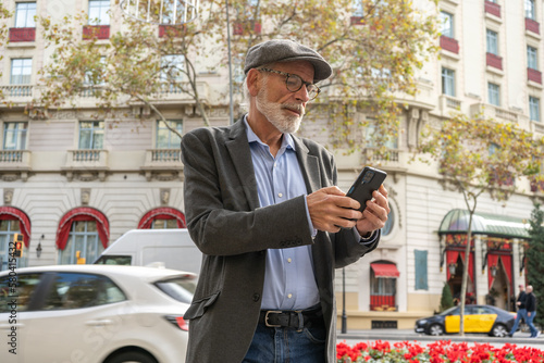 Mature man with gray hair and beard in blue shirt, jacket and cap standing on a street with buildings and texting with smartphone