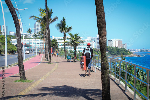 Man with cap and backpack walking on the edge of the beach, next to the bike path, coconut trees and under the blue sky, in the background buildings and apartments