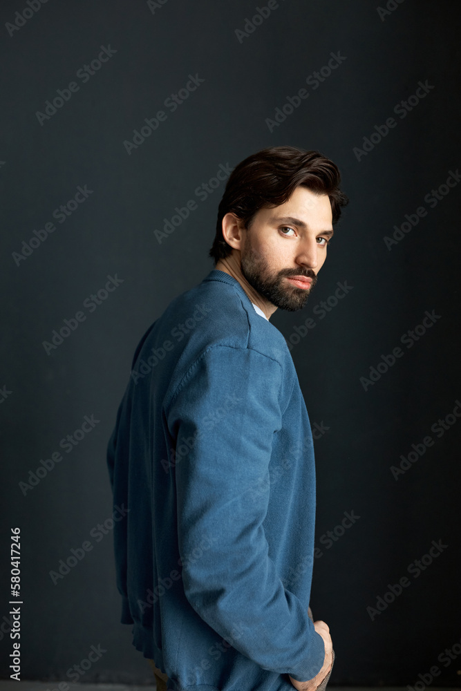 Rare view studio portrait of brutal charming bearded male model in blue cardigan wearing new men collection of warm stylish clothes, turning head at camera, isolated on black . Menswear