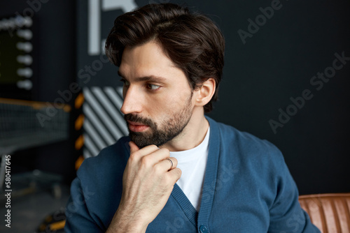 Portrait of young brutal bearded handsome pensive male trader sitting on brown couch with hand under chin, looking aside, wearing wedding ring, planning strategy of trading at stock market