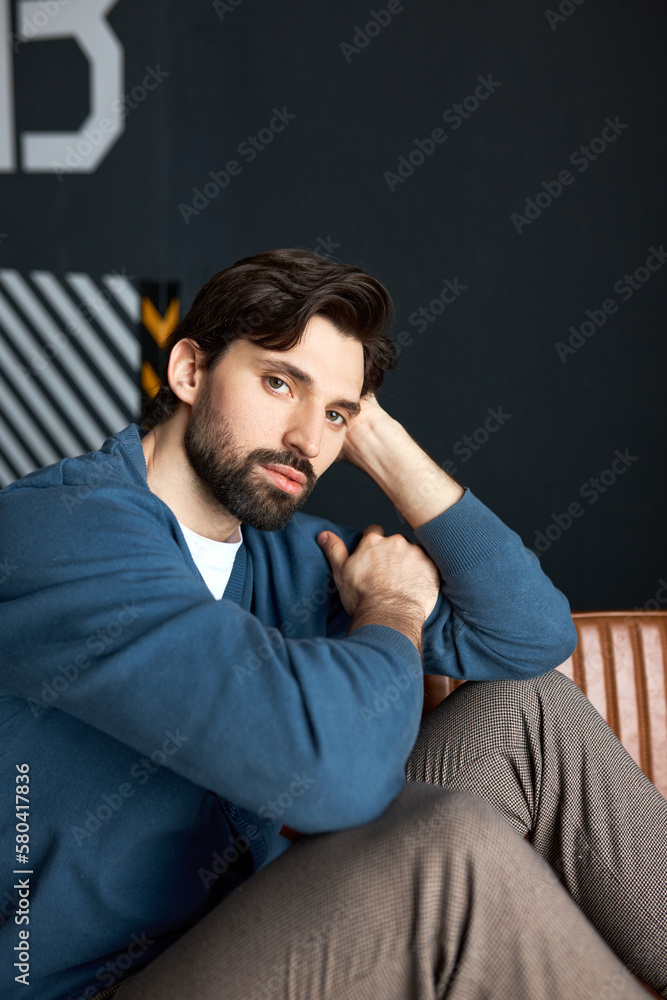 Handsome brutal hipster guy looking at camera with serious confident facial expression, posing in blue cardigan and stylish pants, touching his head, putting elbow on knee. Male fashion Handsome brutal hipster guy looking at camera with serious confident facial expression, posing in blue cardigan and stylish pants, touching his head, putting elbow on knee. Male fashion