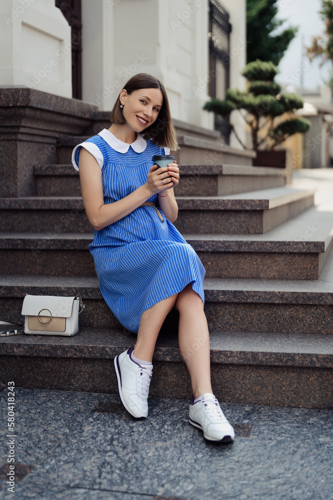 Fototapeta premium Pregnant woman in a blue dress touching her tummy while sitting and resting in the city on the stairs. Hands holding a cup with copy space
