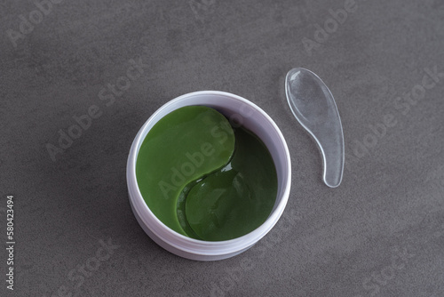 Green eye patches with spatula in a white plastic jar on a grey textured background.