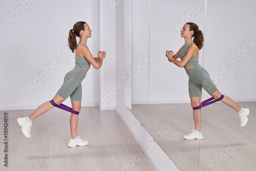 Young healthy woman standing in front of mirror and training with rubber band during training in gym