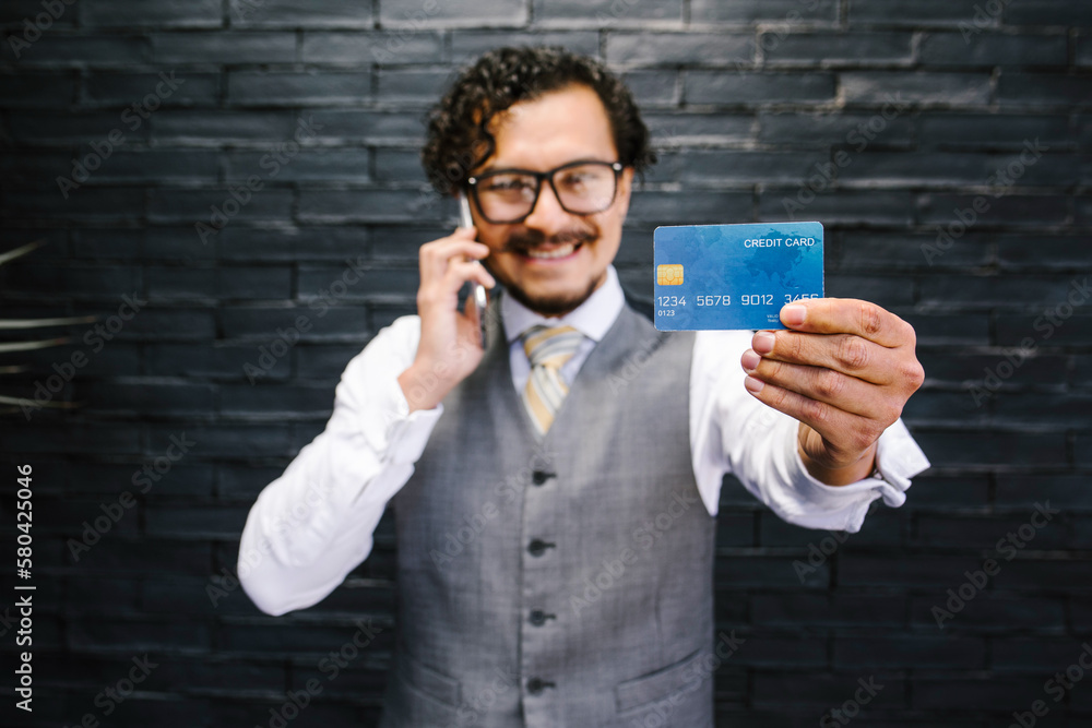 young hispanic businessman holding credit card for shopping and talking by mobile phone in Mexico Latin America young hispanic businessman holding credit card for shopping and talking by mobile phone in Mexico Latin America
