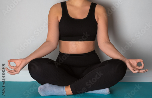 A woman sitting in lotus position on a turquoise yoga mat in a room with light walls.