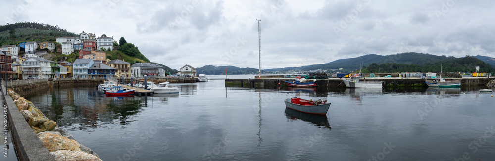 Panoramic view of the village of O Porto do Barqueiro, Galicia, with boats moored at the pier, reflected in the blue water of sea, mountain in the background with houses traveling in summer of 2021