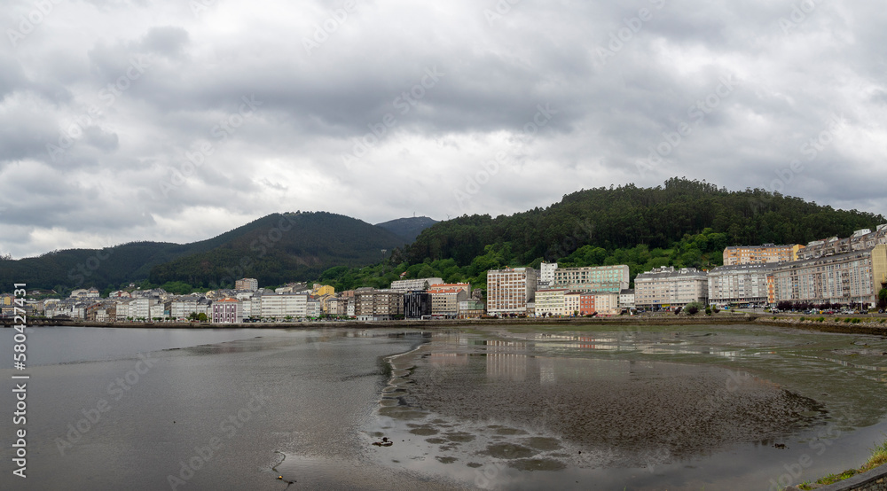 Vistas panorámicas de la ría de Viveiro en Lugo, con marea baja y arena mojada reflejando los edificios de la orilla, montañas verdes y cielo de verano nuboso en España 2021