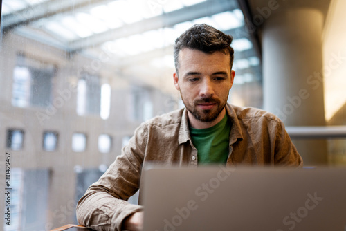 Portrait of pensive, handsome Latin student using laptop computer, Freelancer working in modern office. Attractive copywriter sitting at workplace. Successful business