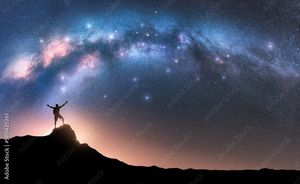 Milky Way and happy man with backpack on mountain peak at night. Silhouette of guy with raised up arm on the hill, sky with stars, yellow light in Nepal. Galaxy. Space landscape with milky way arch