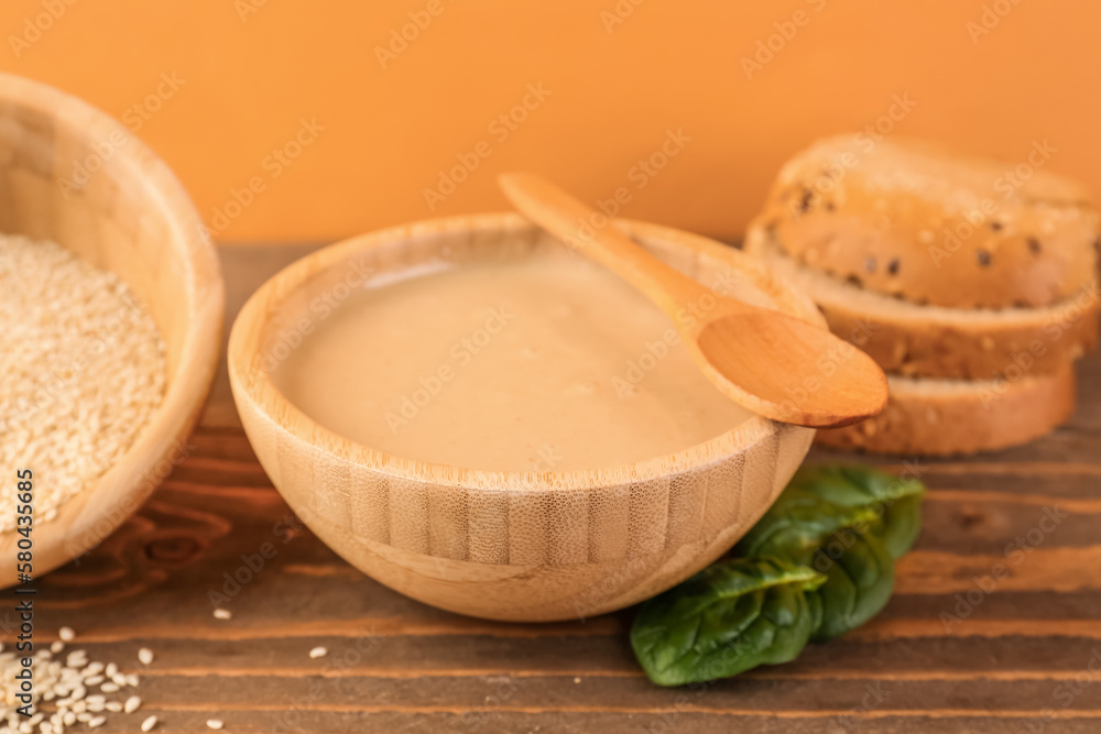 Bowls with tasty tahini, sesame seeds, bread and spinach on wooden table near color wall, closeup Bowls with tasty tahini, sesame seeds, bread and spinach on wooden table near color wall, closeup