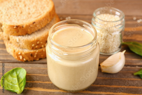 Jars with tasty tahini  sesame seeds  bread and spinach on wooden table  closeup