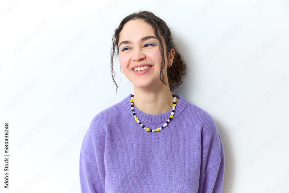 Portrait of cute creative female with wavy hair and ponytail wears vibrant make up on purple eye shadow fancy necklace and purple sweater smiles laughs and looks away isolated over gray background. Portrait of cute creative female with wavy hair and ponytail wears vibrant make up on purple eye shadow fancy necklace and purple sweater smiles laughs and looks away isolated over gray background.