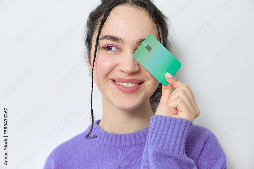 Happy young woman with curly hair and braids smiling looking away while holding a green credit card over her eye. Cheerful woman with curly hair recommending electronic banking and online shopping. Happy young woman with curly hair and braids smiling looking away while holding a green credit card over her eye. Cheerful woman with curly hair recommending electronic banking and online shopping.