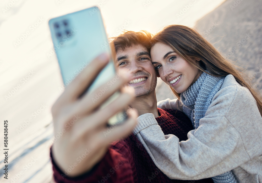 Fototapeta premium Beautiful young couple making photos with mobile phone in a cold winter on the beach.