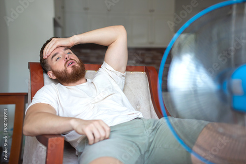 Young bearded man using electric fan at home, sitting on couch cooling off during hot weather, suffering from heat, high temperature