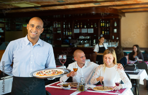 Hospitable latin american waiter standing with serving tray in pizza restaurant hall, welcoming guests photo