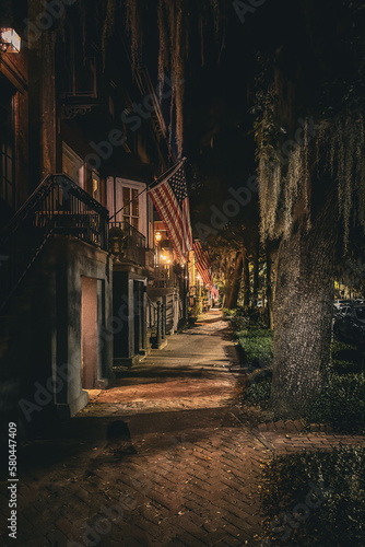 View down old brick-paved street of Savannah, Georgia at night illuminated by gas lamps with a line of American flags - Wide
