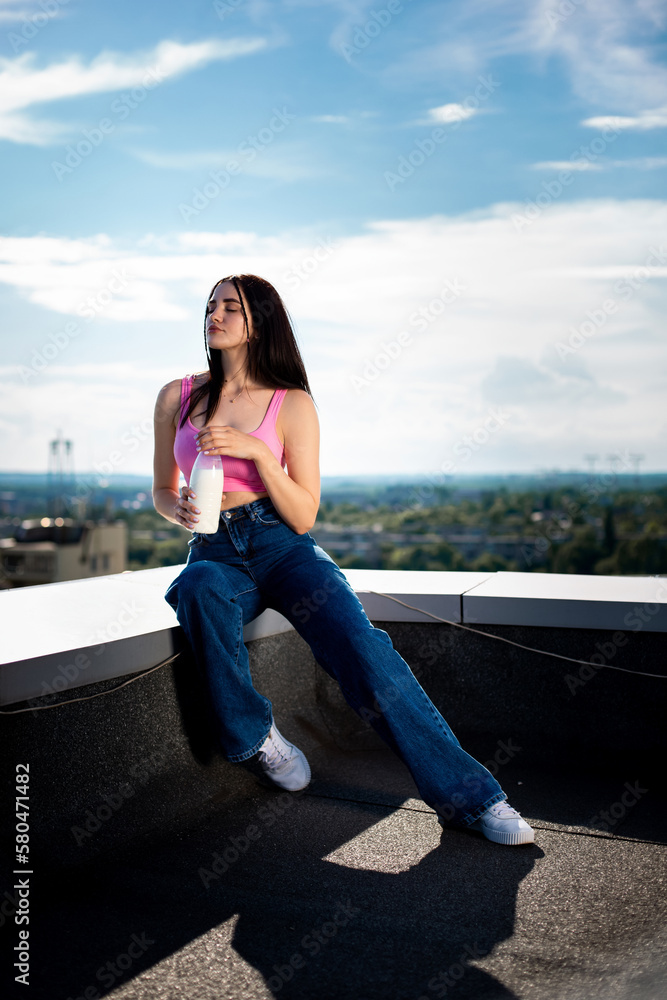 Beautiful sexy slim brunette caucasian girl in pink top and blue jeans sitting on the roof holding a bottle of milk with softly blurred sky background Beautiful sexy slim brunette caucasian girl in pink top and blue jeans sitting on the roof holding a bottle of milk with softly blurred sky background