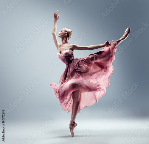 Ballerina in Pink Chiffon Dress jumping Split. Ballet Dancer in Silk Gown Pointe Shoes. Graceful Woman in Tutu Skirt dancing over Gray Background