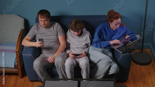 The family sits on the couch without talking to each other. Addicted to gadgets, family using mobile phone, laptop ignoring each other at home. Internet obsession concept photo
