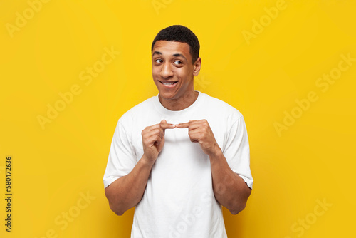 young bashful african american guy in white t-shirt is shy on yellow isolated background, embarrassed man
