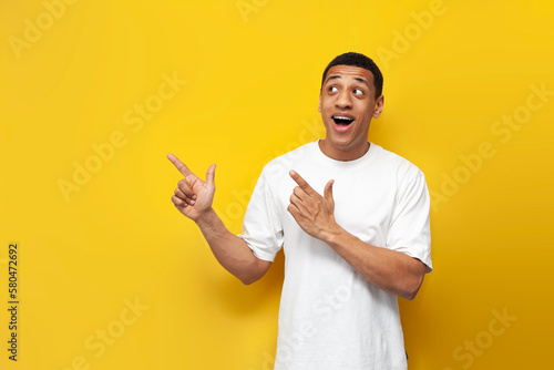 young african american guy in white t-shirt shows his hands to the side on yellow isolated background