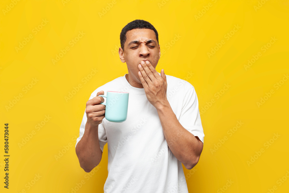 young tired african american guy in white t-shirt holds cup of coffee and yawns on yellow isolated background young tired african american guy in white t-shirt holds cup of coffee and yawns on yellow isolated background