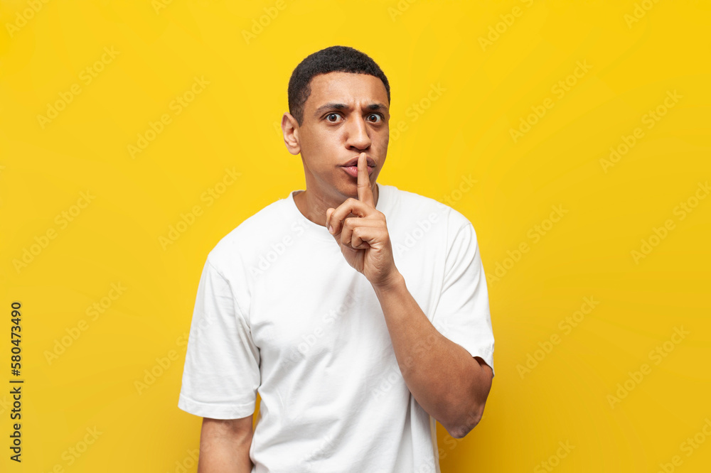 young african american guy in white t-shirt hides secret and shows gesture of silence, man covers his mouth young african american guy in white t-shirt hides secret and shows gesture of silence, man covers his mouth