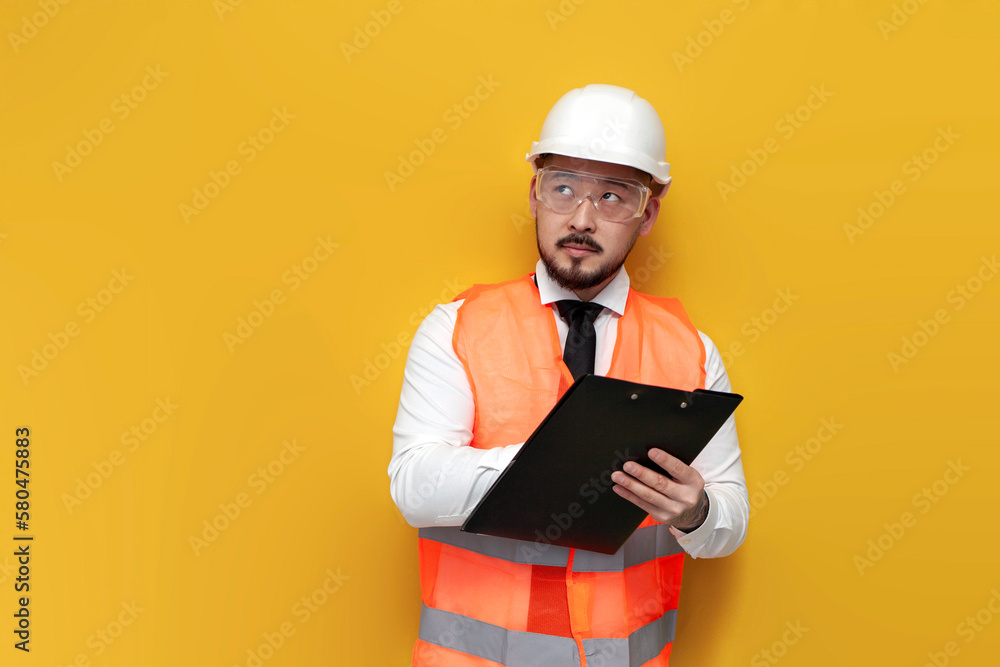 asian civil engineer in uniform writes plan on paper and looks away on yellow isolated background asian civil engineer in uniform writes plan on paper and looks away on yellow isolated background