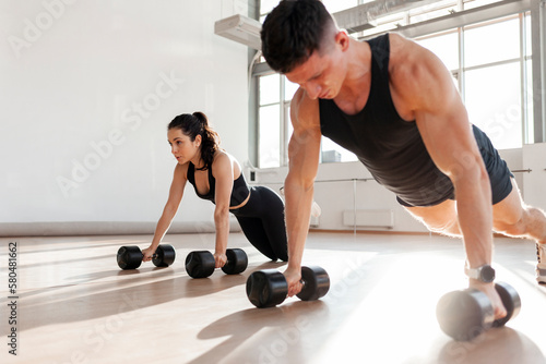 Fototapeta Naklejka Na Ścianę i Meble -  athletic couple in sportswear in training do push-ups with dumbbells in the fitness room in the morning