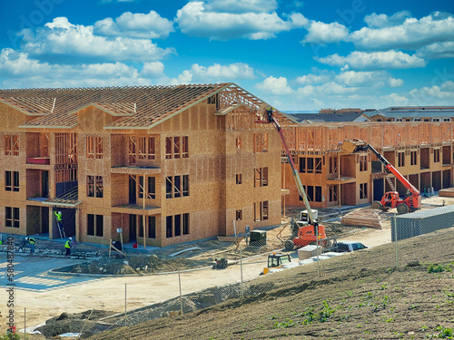 Hard working team constructing multi-family housing apartments: Construction workers utilizing heavy duty equipment, trucks, and articulating boom lifts to finish exterior walls and roof with plywood