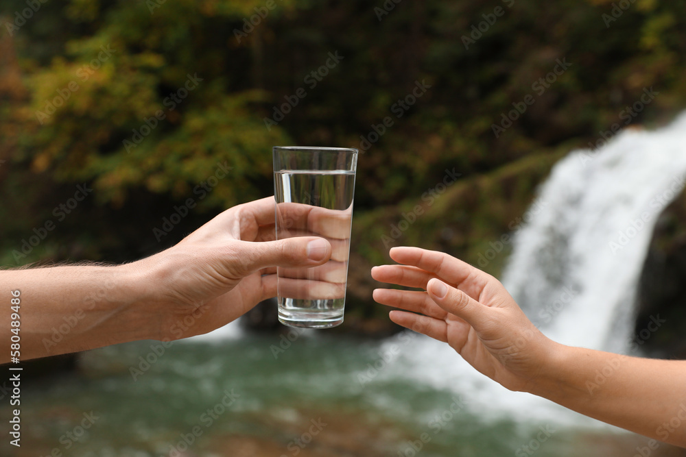 Man giving woman glass of fresh water near waterfall, closeup Man giving woman glass of fresh water near waterfall, closeup