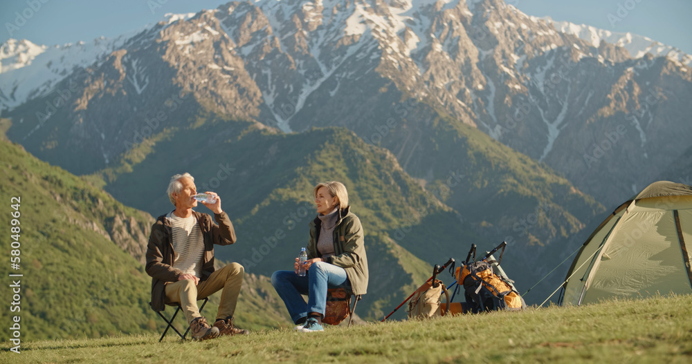 Old caucasian couple taking a rest on top of mountain, sitting and rehydrating while enjoying the view, talking. Mature people spending time together after retirement travelling - tourism, pension - obrazy, fototapety, plakaty 