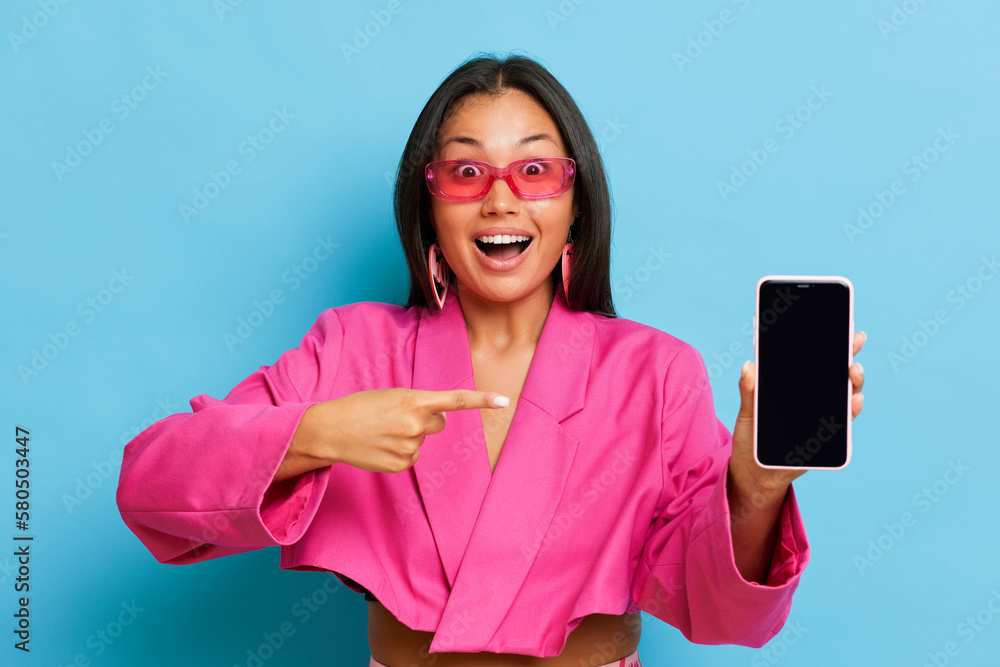 Naklejka premium Young woman in pink sunglasses and jacket points finger at her phone in surprise, girl standing isolated on blue background, big discounts in favorite market, copy space, high quality photo