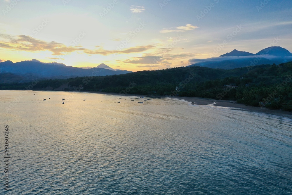 Panoramic drone shot sideways along the beach of Ende on Flores with a golden sunset behind hills.
