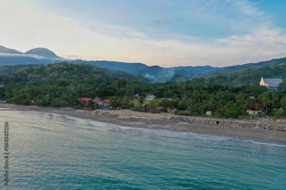 Panoramic drone shot along the beach in Ende on Flores, in the foreground the turquoise sea, in the background the beach and a rainforest area.