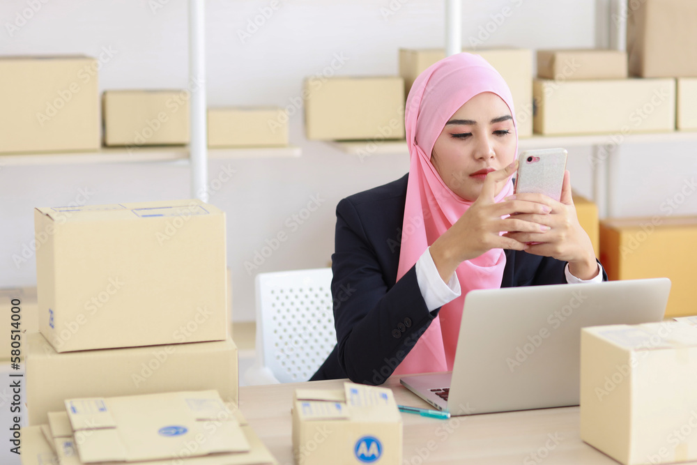 Religious asian woman in muslim dress sitting and using smart mobile phone and talking to customer with happy face and business package SME box delivery background. Religious asian woman in muslim dress sitting and using smart mobile phone and talking to customer with happy face and business package SME box delivery background.