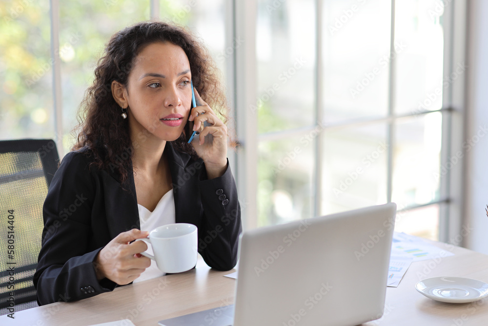 Attractive professional latin female employee worker sitting, using laptop computer with paperwork at home workplace. Businesswoman talking on smart mobile phone and communicate with client. Attractive professional latin female employee worker sitting, using laptop computer with paperwork at home workplace. Businesswoman talking on smart mobile phone and communicate with client.